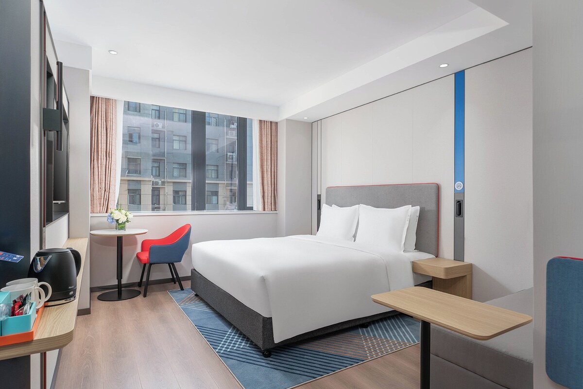 A modern hotel room with a large bed, a desk with a chair, a coffee machine, and a view of the city through the window.