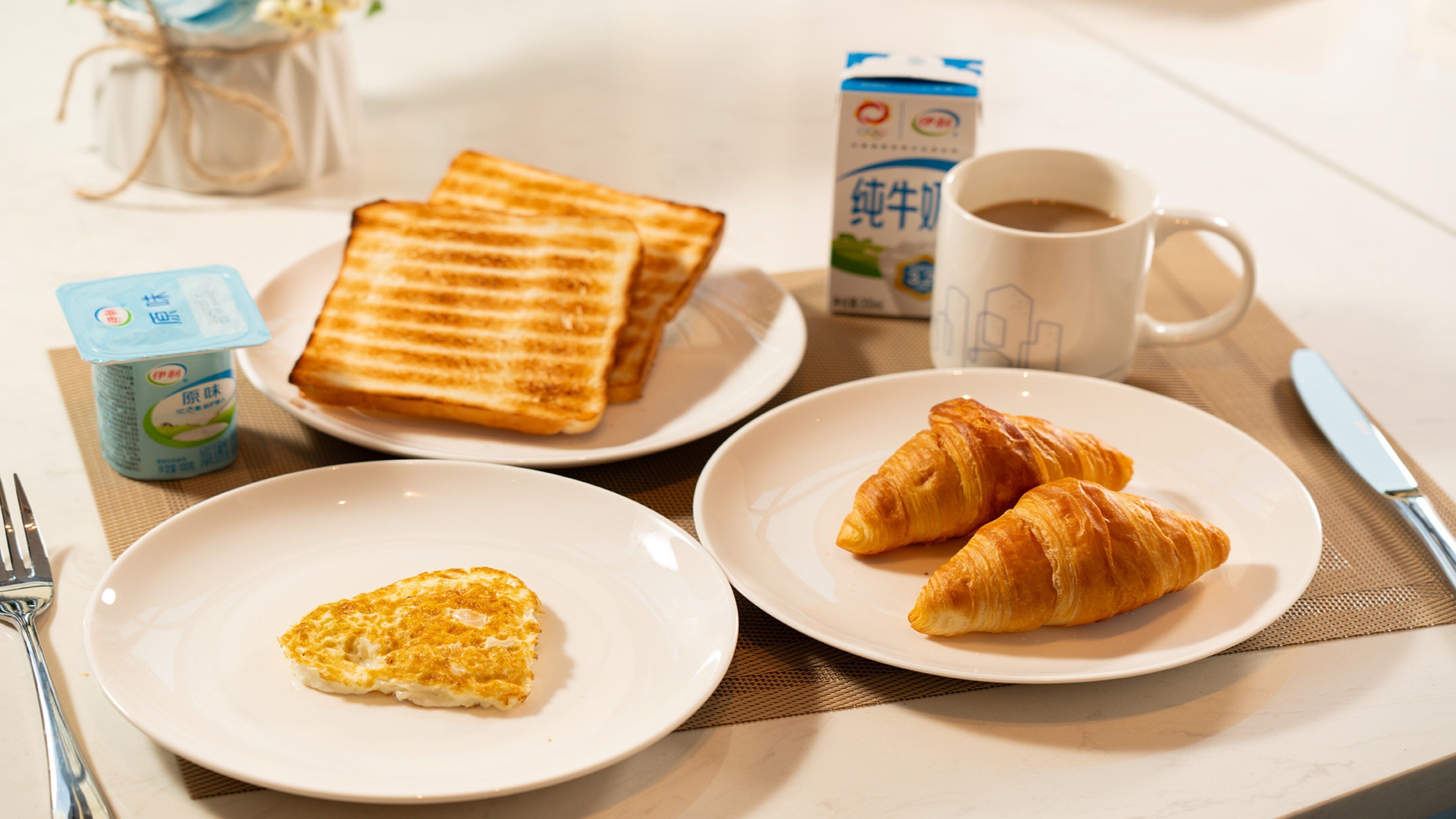 A breakfast setting with toast, croissants, a cup of coffee, and a yogurt container.