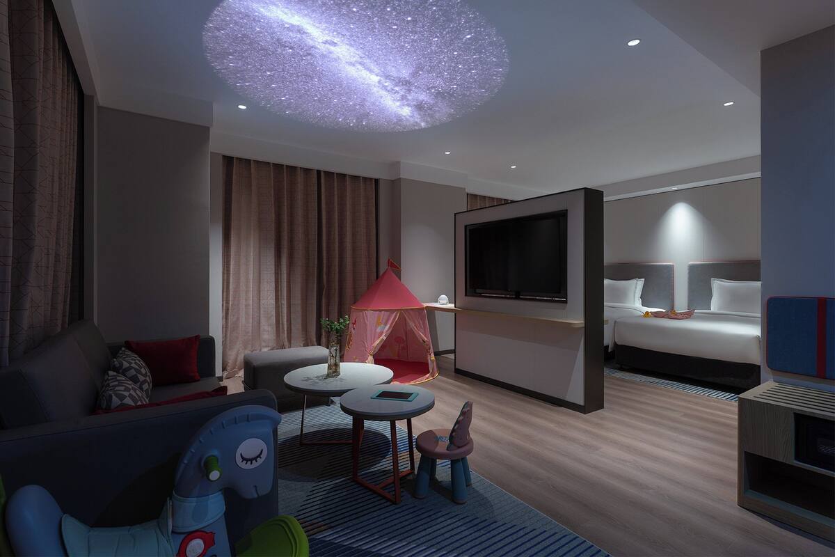 Una habitación de hotel moderna con una cama grande, un televisor, un proyector de techo que muestra un cielo estrellado, una pequeña zona de juegos con una tienda de campaña, y un área de descanso con un sofá y una mesa de centro.