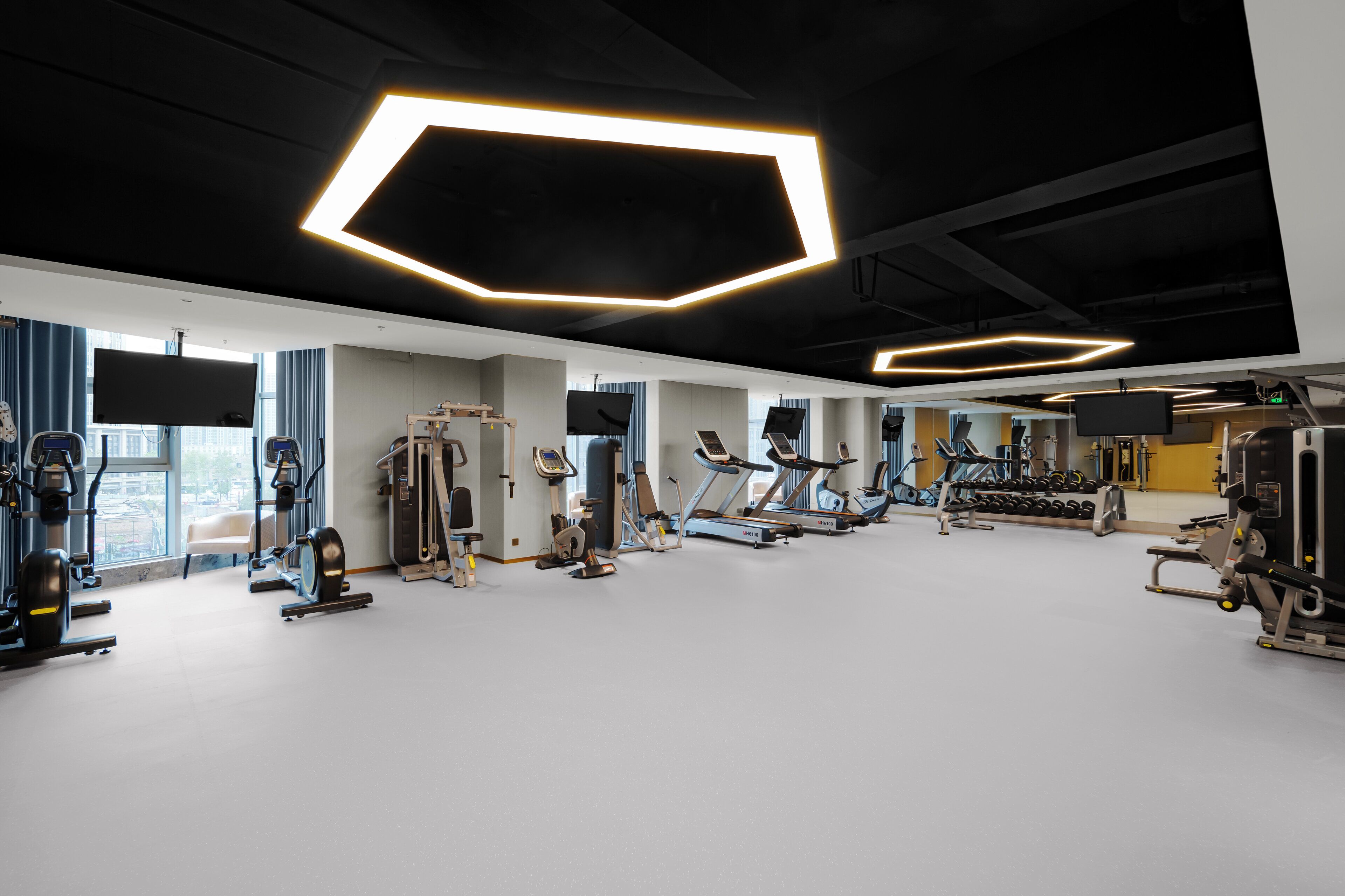 Un gimnasio moderno con varios equipos de ejercicio, ventanas amplias y luminarias geométricas en el techo.