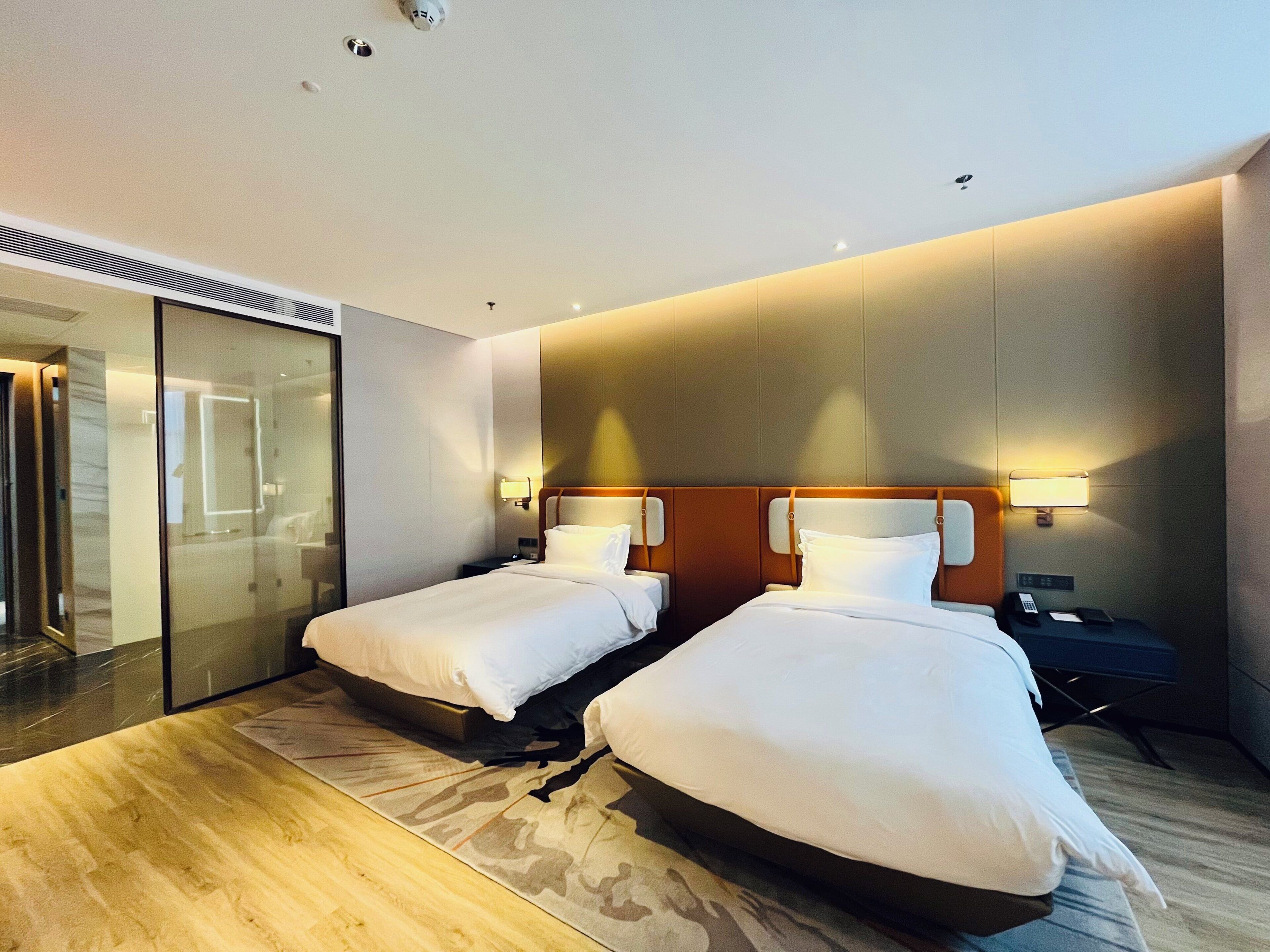 Habitación de hotel con dos camas, una mampara de vidrio, piso de madera y una lámpara fijada a la pared.