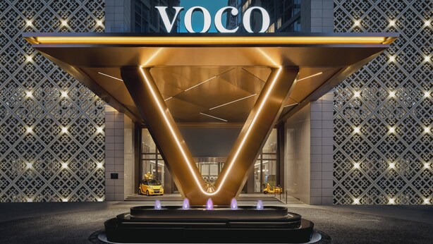 Une entrée d'hôtel moderne, avec une enseigne « VOCO » bien visible, un auvent en verre aux formes géométriques et une façade à motifs.