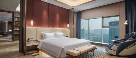Una habitación de hotel moderna con una cama grande, zona de estar con sillón de masaje, vistas a la ciudad y techo bien iluminado.