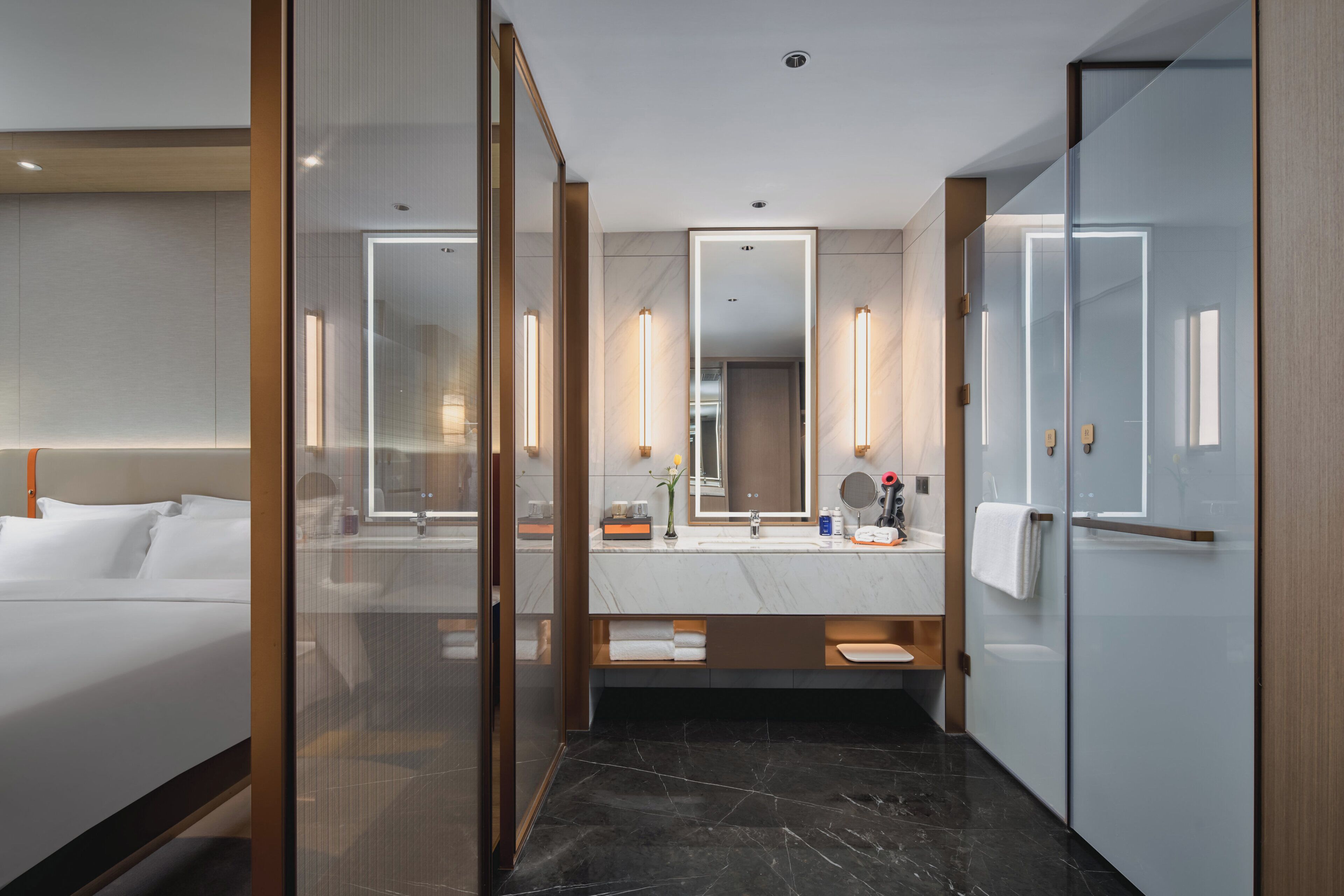 A modern hotel room with a large bed, a glass-enclosed shower, a marble-topped vanity, and a large mirror.