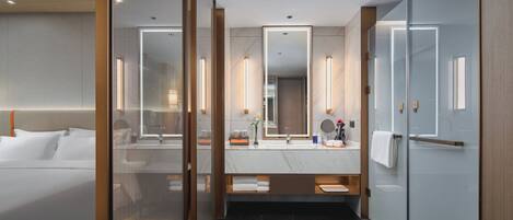 A modern hotel room with a large bed, a glass-enclosed shower, a marble-topped vanity, and a large mirror.