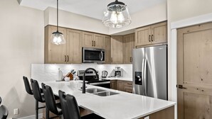 A modern kitchen with wooden cabinets, stainless steel appliances, a white countertop, and black bar stools.