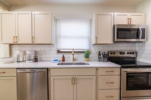 Una cocina con armarios blancos, lavavajillas de acero inoxidable, microondas, horno y una ventana con persianas.