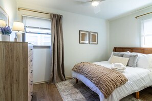 Un dormitorio con cama, mesita de noche, lámpara, ventana con cortinas y dos cuadros en la pared.