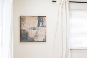 Una habitación con un cuadro abstracto enmarcado en la pared, una ventana con persianas y una cortina medio bajada.