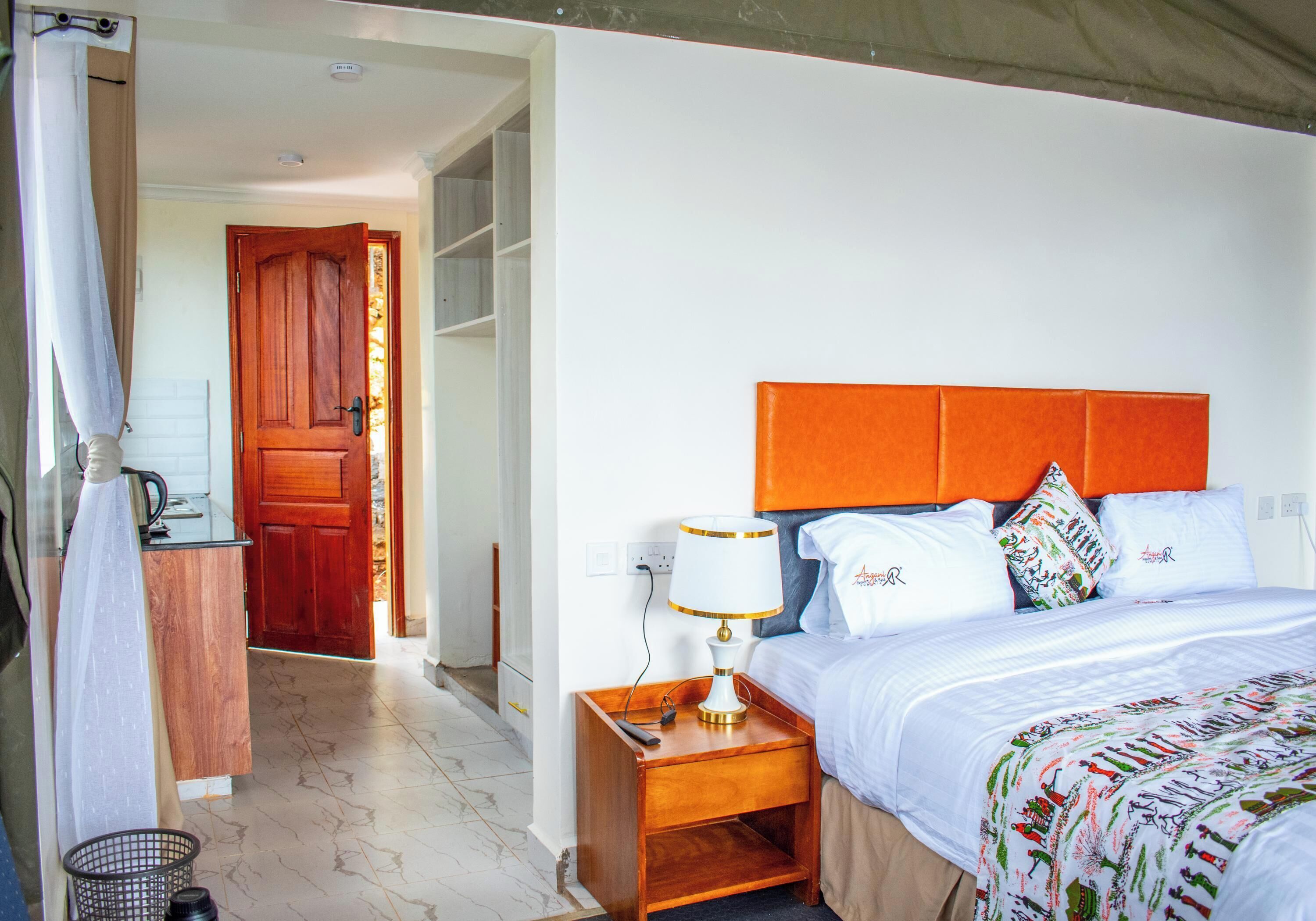 A hotel room with a bed, a nightstand with a lamp, a wooden door, and a white wall.