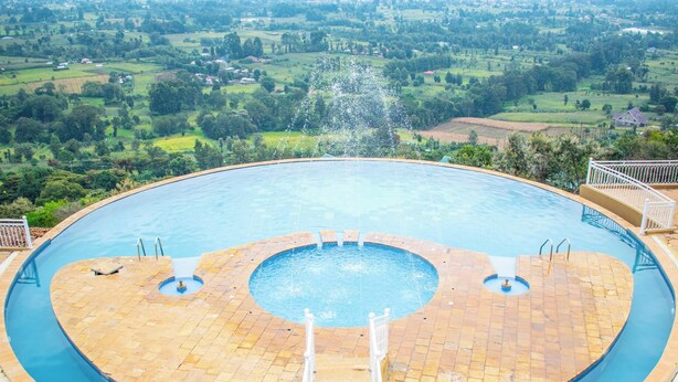 Uma grande piscina circular com uma fonte central, rodeada por um deck pavimentado e um guarda-corpo branco.