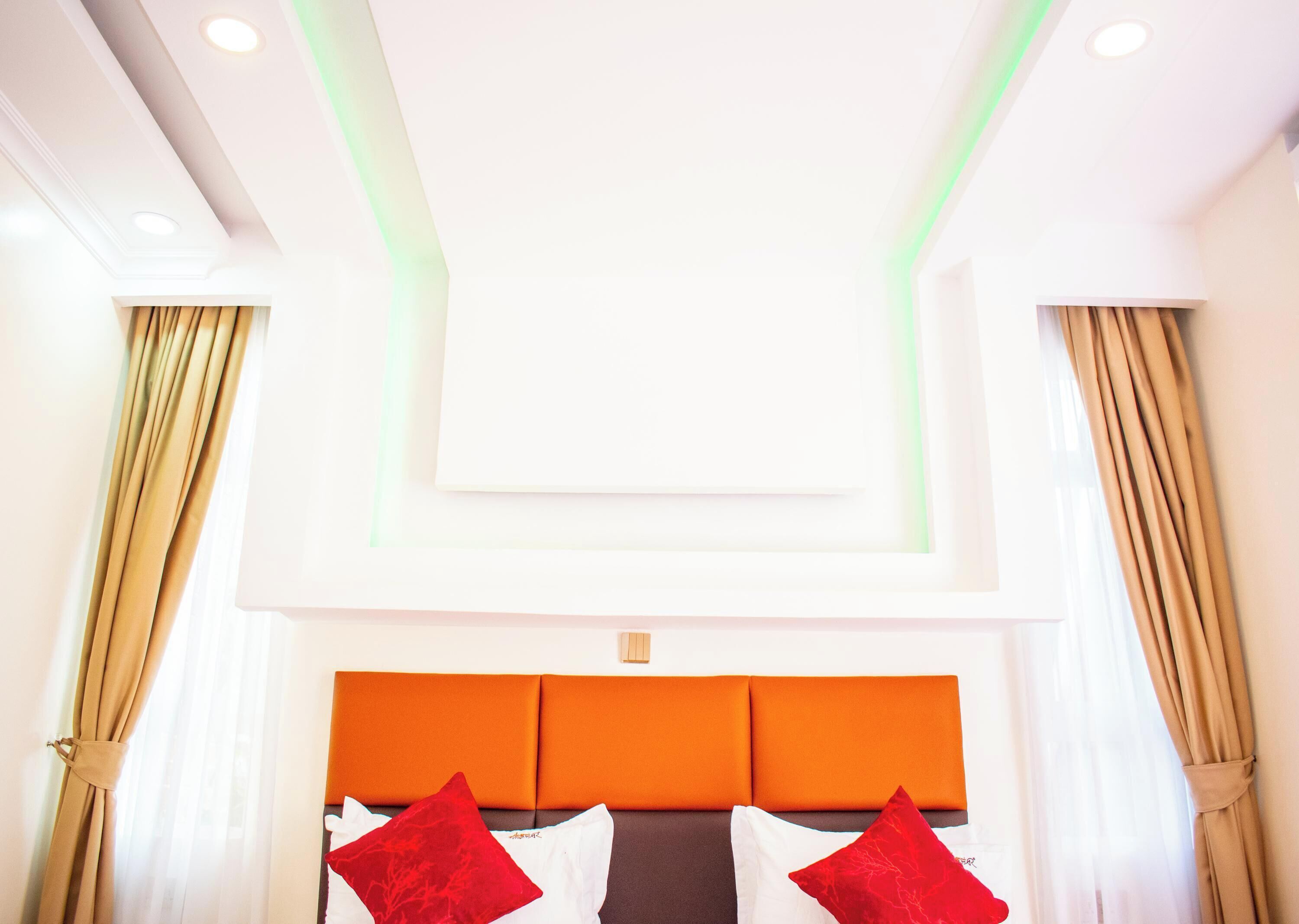 Habitación de hotel con diseño moderno, que presenta una cama grande con cabecera naranja, dos almohadas rojas y cortinas color beige.