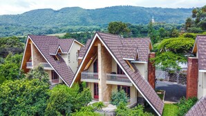 Uma casa moderna com um design triangular único no telhado, rodeada por vegetação exuberante e colinas ao fundo.