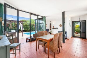 A spacious living area with a dining table and chairs, a pool visible through large glass doors, and a view of lush greenery outside. - ANANA Binisafua (Sant Lluis)