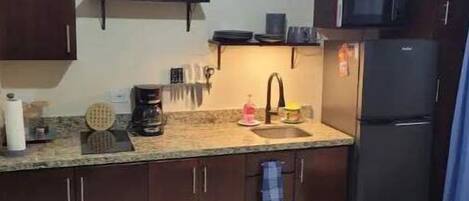 A compact kitchen with dark wooden cabinets, a microwave, a refrigerator, and a clock on the wall.