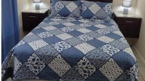 A bed with a patterned quilt, two bedside tables with lamps, a wall-mounted artwork, and a blue curtain. - Craig's Coco Condo (Liberia)