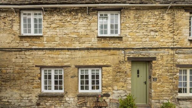 A stone building with a green door, two windows with shutters, and a small outdoor seating area with a table and chairs. - Beautiful Historic High Street Cottage in Burford (Burford)