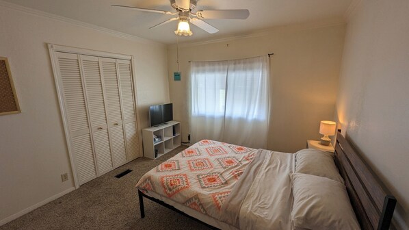 Un dormitorio con cama, televisor sobre un mueble, ventilador de techo y ventana con cortinas.