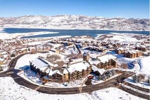 Estância coberta de neve com um edifício central rodeado por quartos de hóspedes, um lago e montanhas ao fundo.
