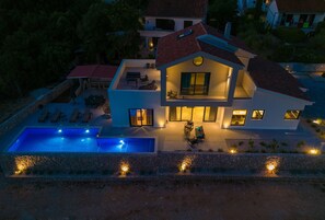 A modern two-story house with a swimming pool, outdoor seating, and a well-lit patio area.