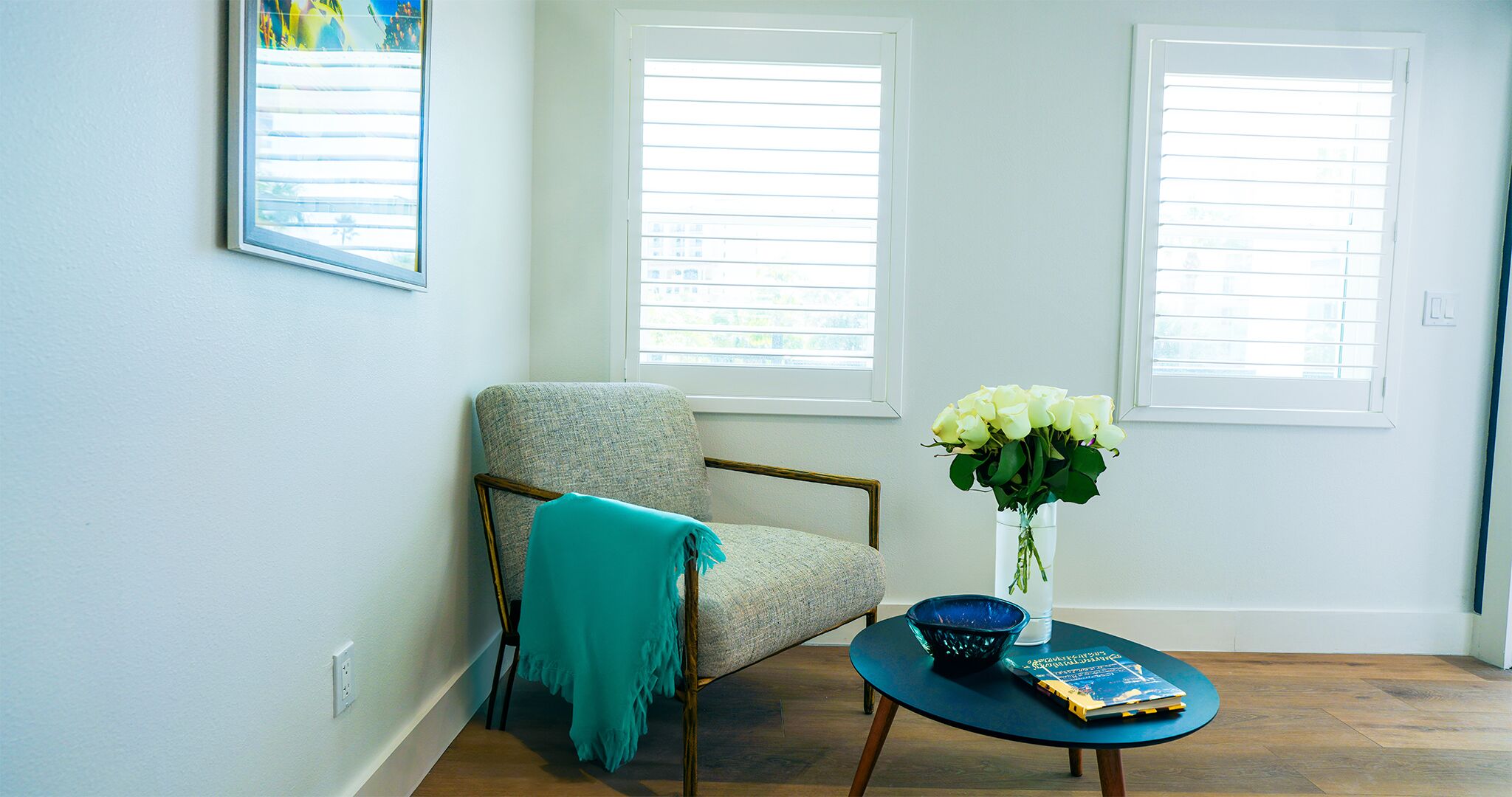 A room with a light-colored armchair, a small table with a book and a vase of flowers, and two windows with blinds.