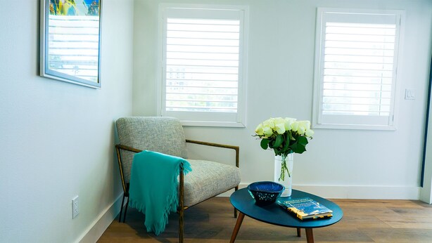 Une pièce avec un fauteuil de couleur claire, une petite table sur laquelle se trouvent un livre et un vase de fleurs, et deux fenêtres équipées de stores.