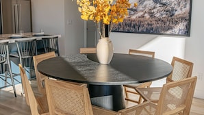 A modern dining area with a round table, six wooden chairs, a vase with yellow flowers, and a large landscape painting on the wall. - Ski&Tee Mountain Chalet: modern retreat w/ golf simulator, hot tub, & game room (Park City)