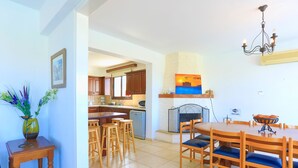 A spacious dining area with a wooden table and chairs, a kitchen in the background, and a decorative painting above the fireplace. - 4 Bedroom Villa, Private Pool, Latchi, Cyprus (Latchi)