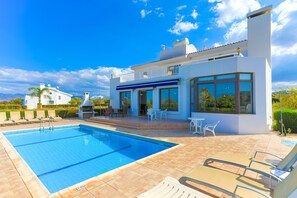 A modern poolside area with a clear blue pool, outdoor seating, and a white building with large windows. - 4 Bedroom Villa, Private Pool, Latchi, Cyprus (Latchi)