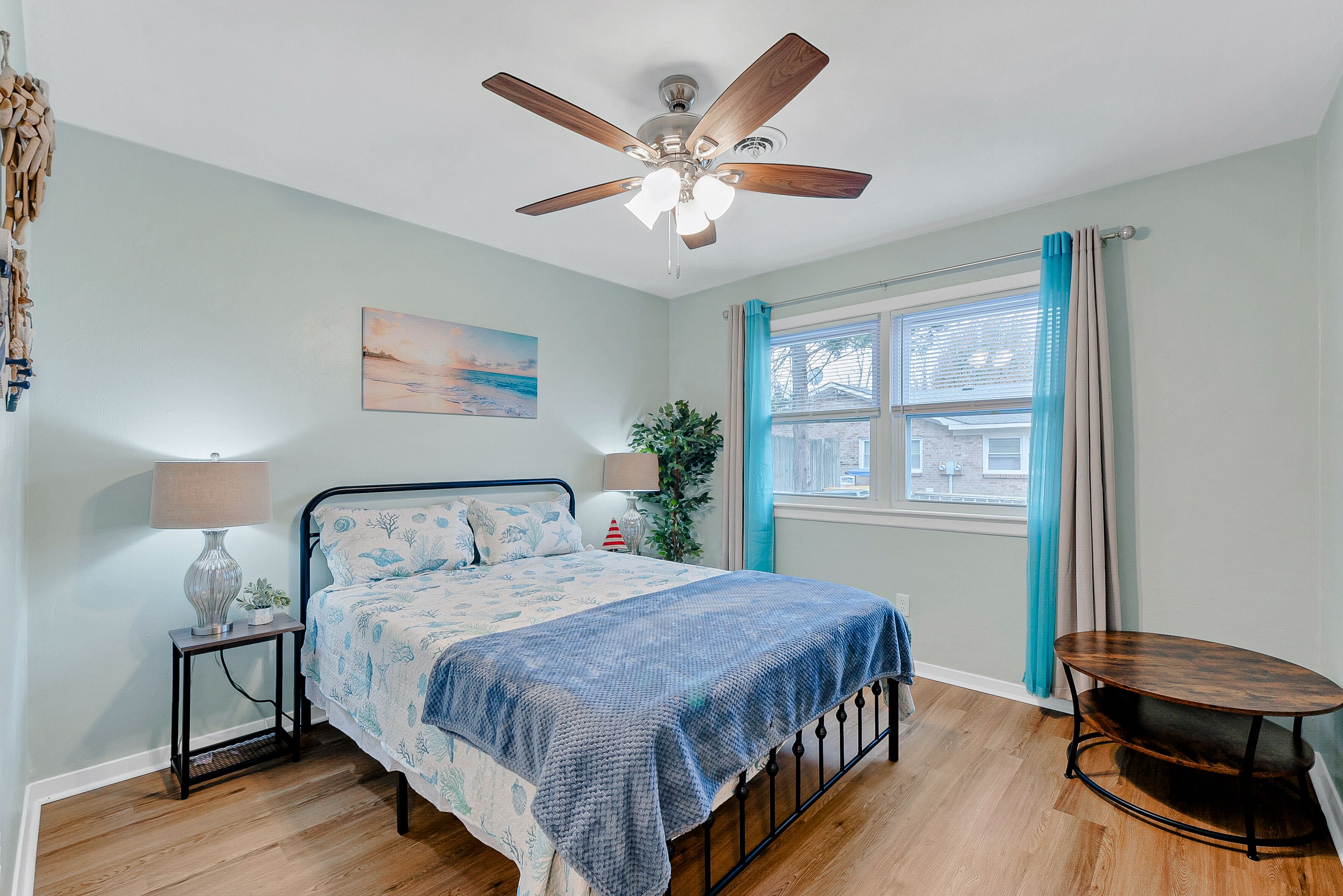 Une chambre à coucher avec un lit, un ventilateur de plafond, une fenêtre avec des rideaux, une table de chevet avec une lampe et une petite plante.