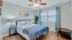 Quarto com cama, ventoinha no teto, janela com cortinas, mesa de cabeceira com abajur e uma pequena planta.