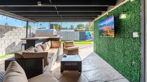 An outdoor patio with wicker furniture, a barbecue grill, and a green wall with a cityscape painting. - 4 Bed Mid Century Modern w/ Pool, Putting Green, Wood Fired Pizza Oven, Sleeps 9 (Phoenix)