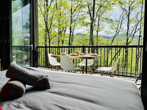 Une chambre avec un grand lit, un balcon avec une table et des chaises, et une vue sur les arbres et les montagnes.