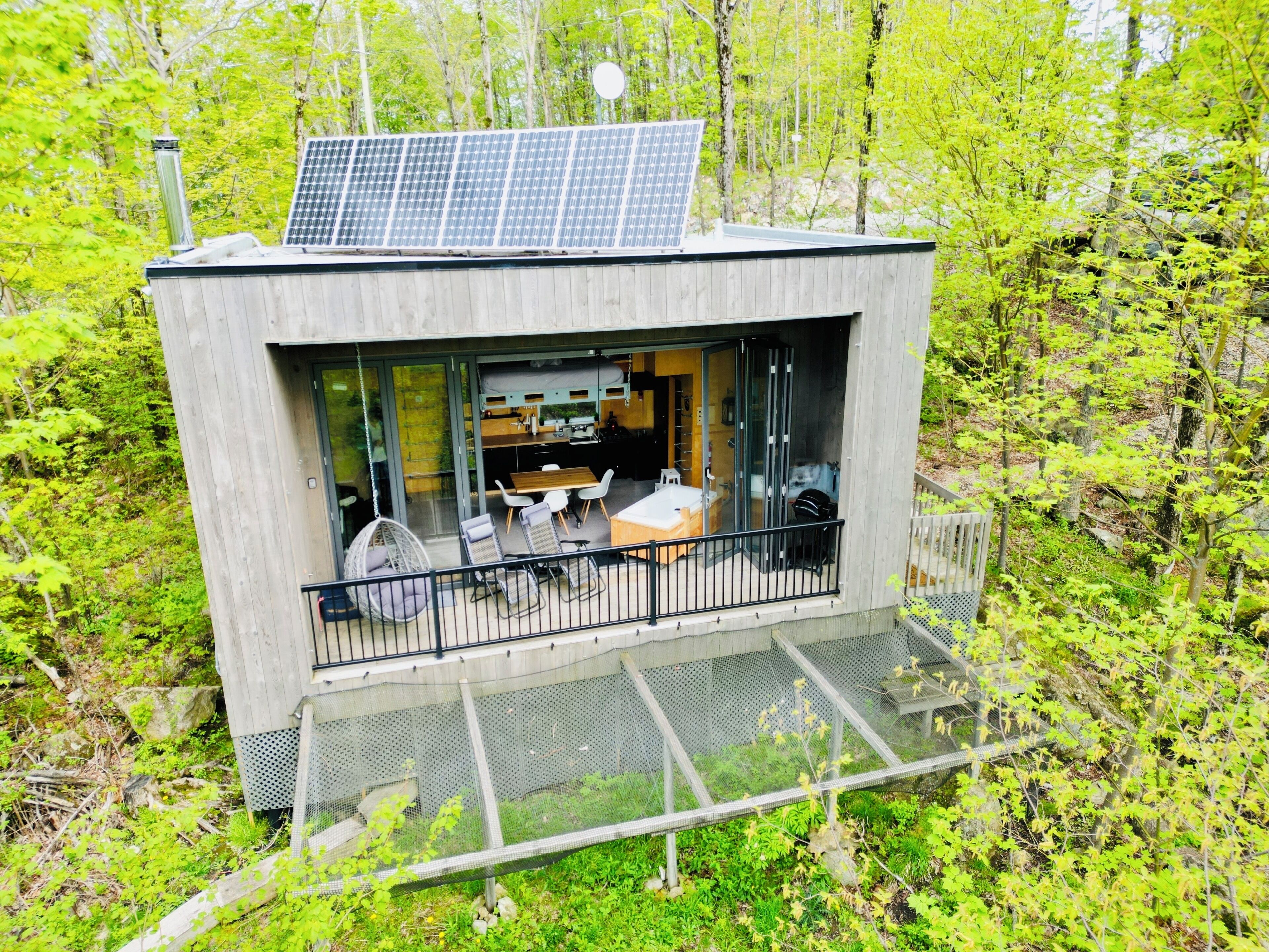 Une cabane moderne dotée d’un balcon, de panneaux solaires et offrant une vue sur la forêt environnante.