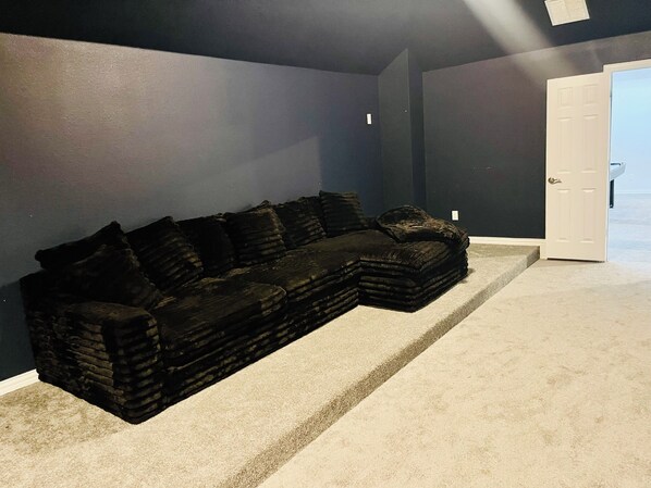 Ein dunkelgraues Ecksofa vor einer dunklen Wand in einem Raum mit Teppichboden und einer weißen Tür.
