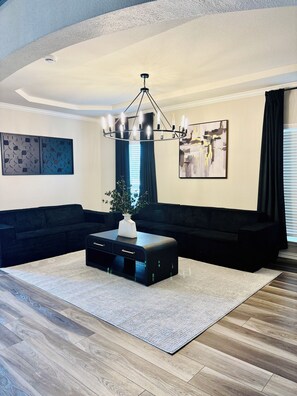 Een moderne woonkamer met een zwart hoekbank, een salontafel met lades, een kroonluchter en een groot abstract schilderij aan de muur.