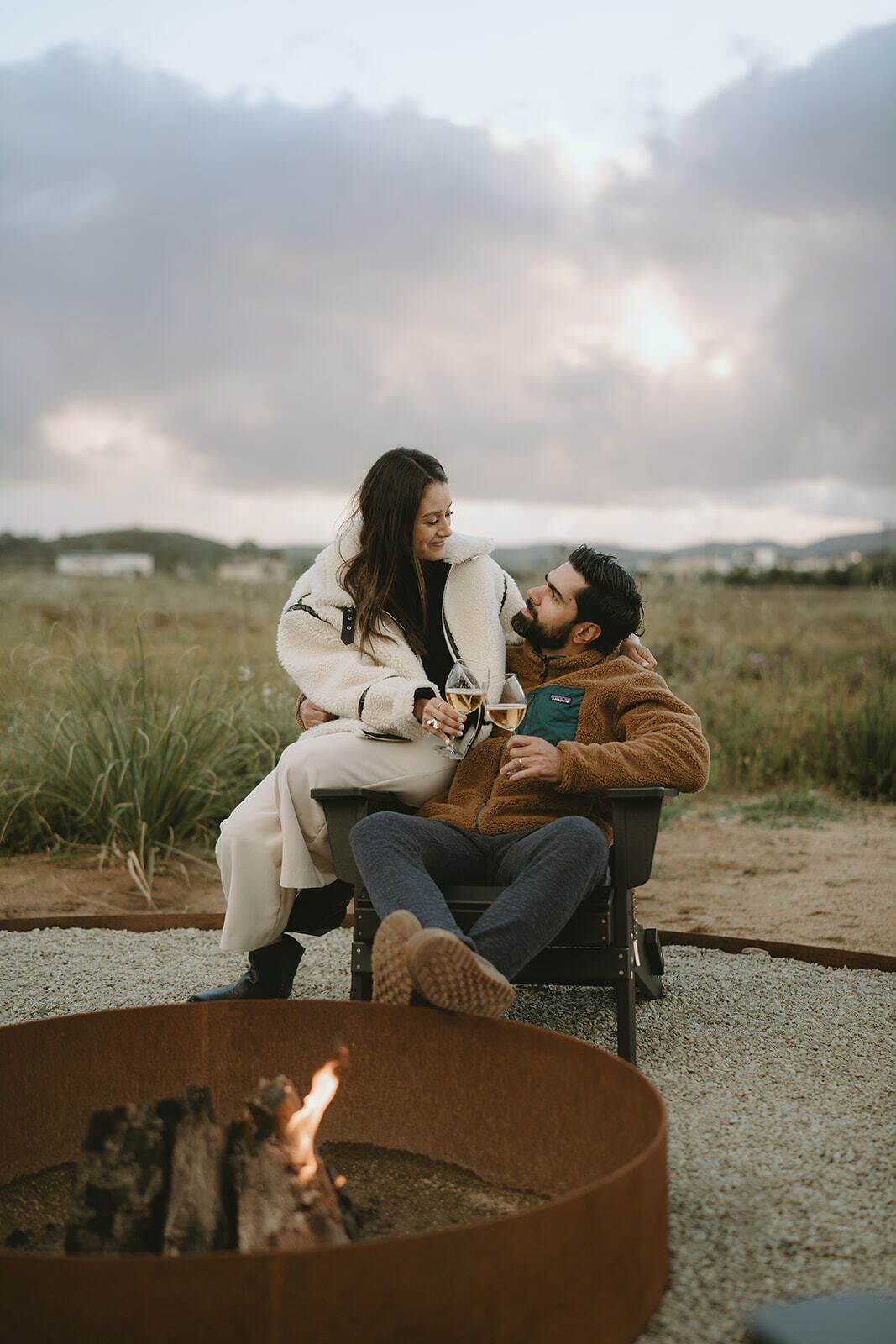 Two people sitting by a fire pit in an outdoor setting with a grassy field and hills in the background.