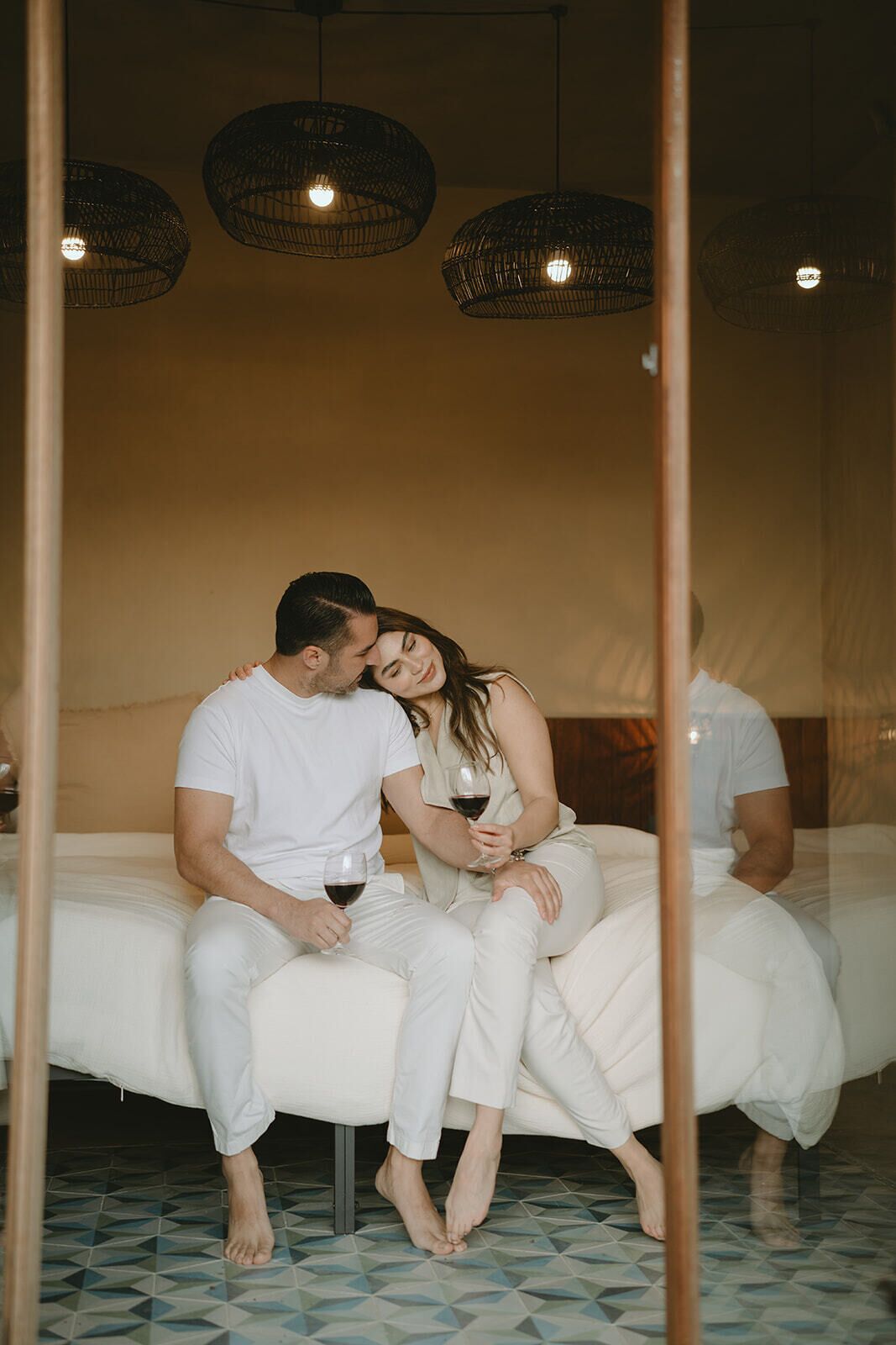 Um casal sentado em uma cama em quarto com chão de azulejos e três luminárias de vime penduradas.