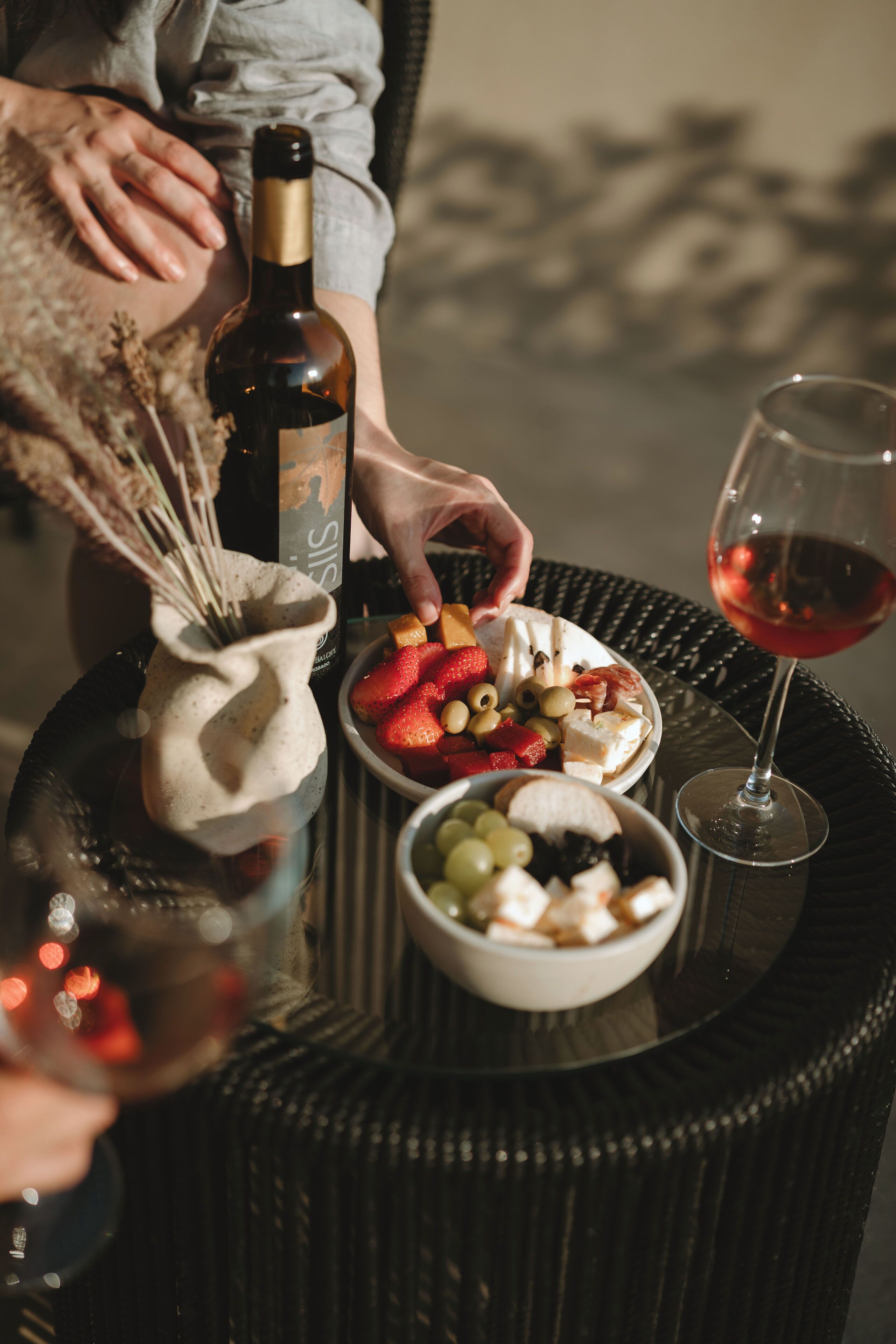 A wicker table with a bottle of wine, a glass of red wine, and bowls of fruit and cheese.