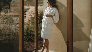 A person in a white robe leaning against a glass wall with a view of a natural landscape. - EQU Hotel Boutique (Valle de Guadalupe)