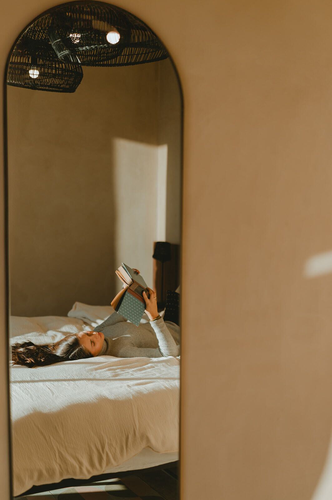 A person lying on a bed reading a book in a room with a large mirror reflecting the bed and a ceiling light fixture.