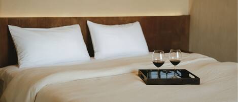 A neatly made bed with white linens and pillows, a tray with two wine glasses and a bottle, and a wooden headboard.