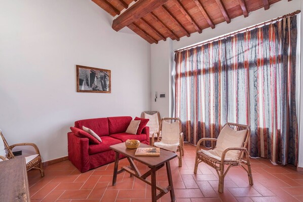 A cozy living room with a red sofa, wicker chairs, a wooden coffee table, and a large window with striped curtains. - Le Masse - Greve in Chianti, Toscana (Greve In Chianti)