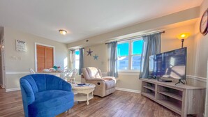 A living room with a blue armchair, a beige sofa, a white armchair, a television, and a window with curtains.