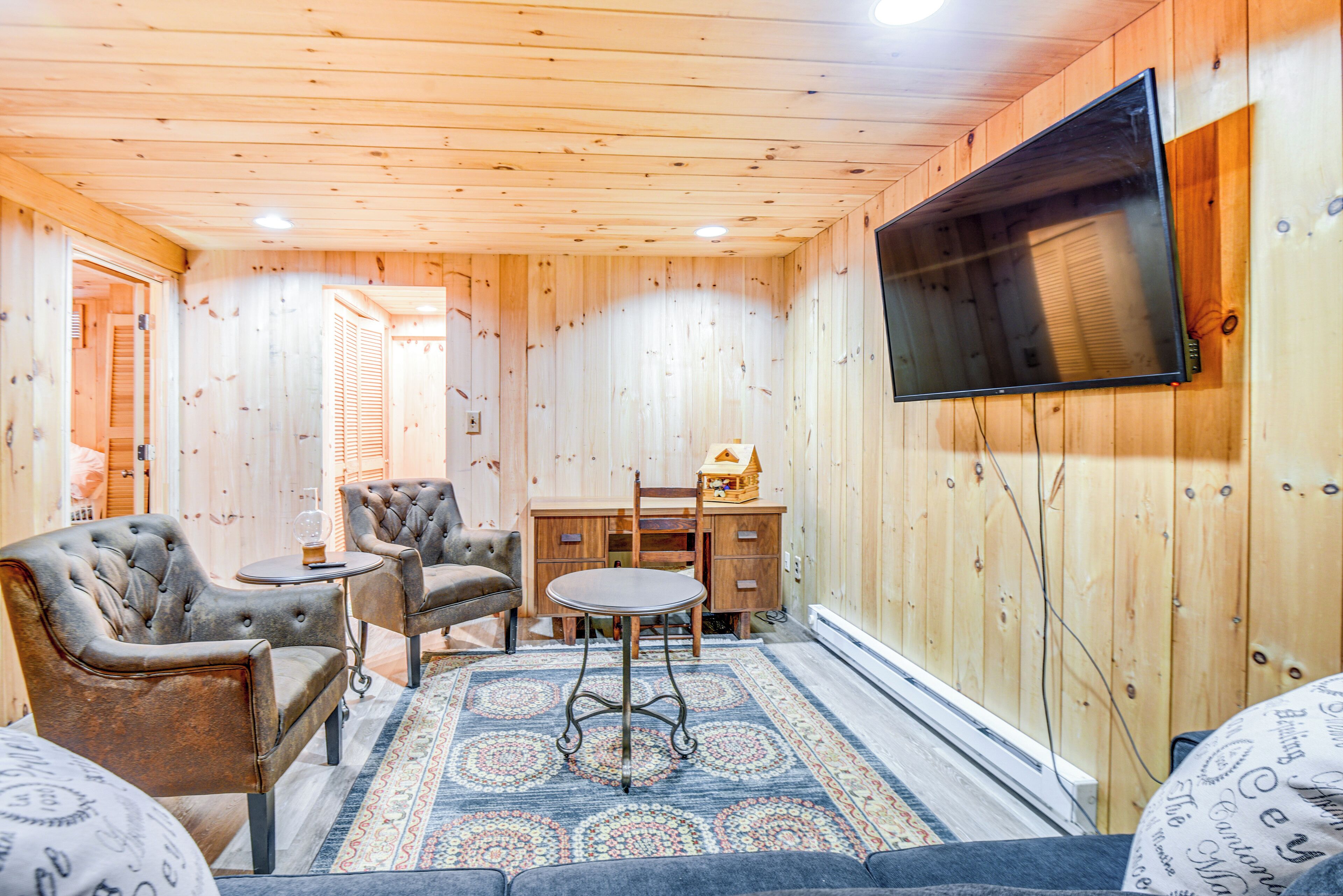 A wooden-paneled room with a television mounted on the wall, two armchairs, a small table, and a patterned rug.
