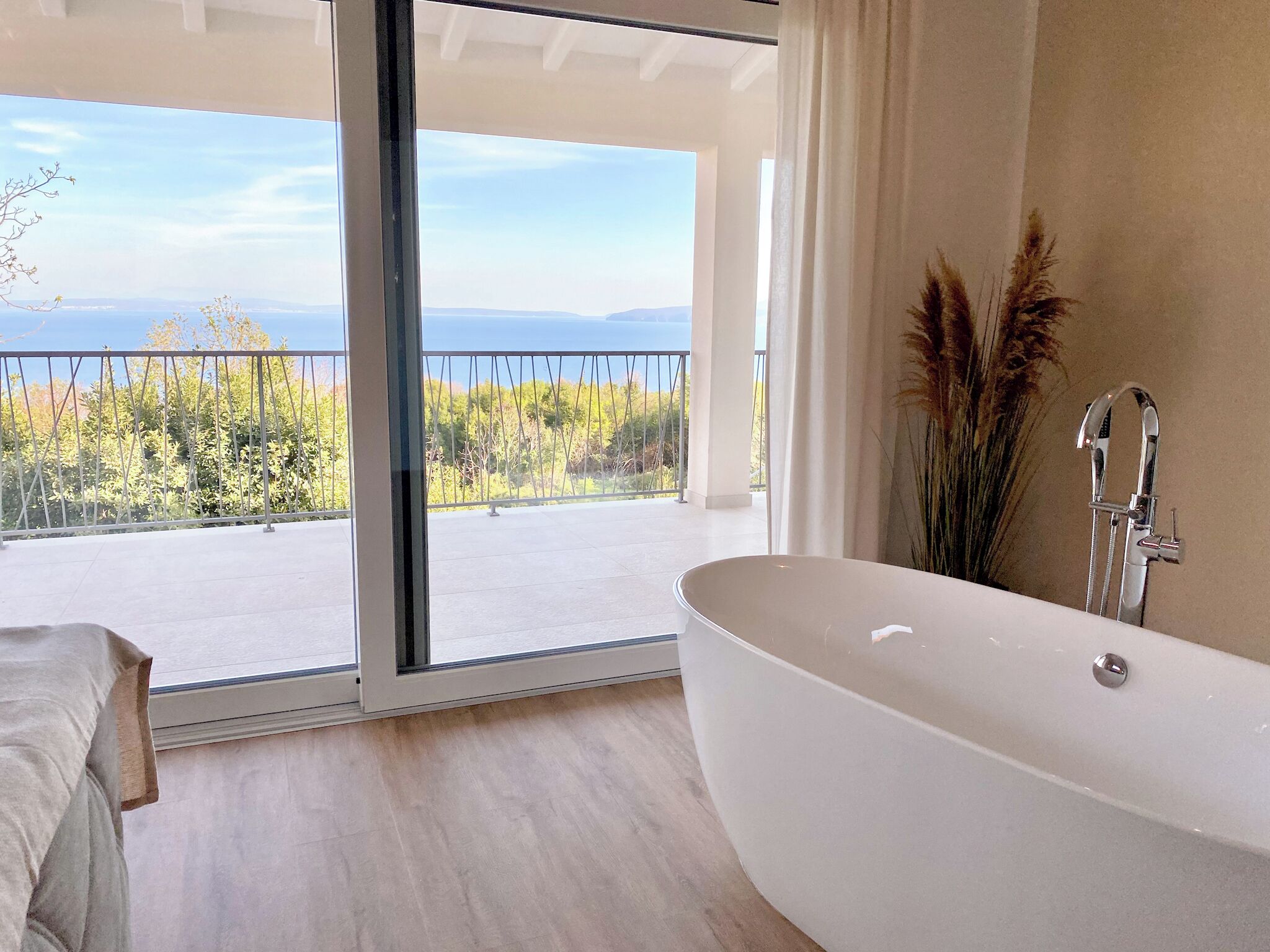 A modern bathroom with a freestanding bathtub, a large window offering a view of trees and a distant landscape, and a shower with a handheld showerhead.