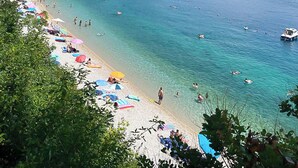 Ein Strand mit klarem, türkisblauem Wasser, Menschen beim Schwimmen und Sonnenbaden, und ein bewaldeter Hügel im Hintergrund.