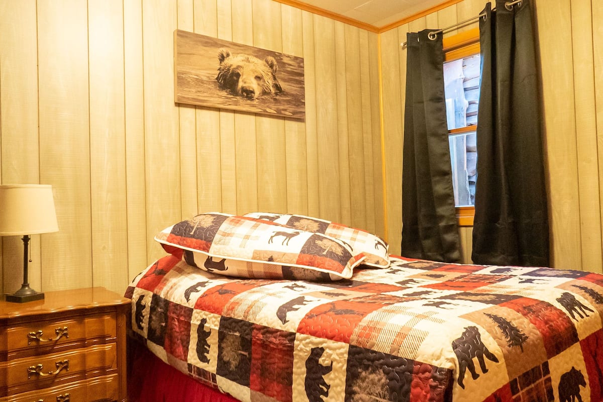 A bedroom with a wooden paneled wall, a bed with a quilt featuring animal prints, a nightstand with a lamp, and a window with curtains.