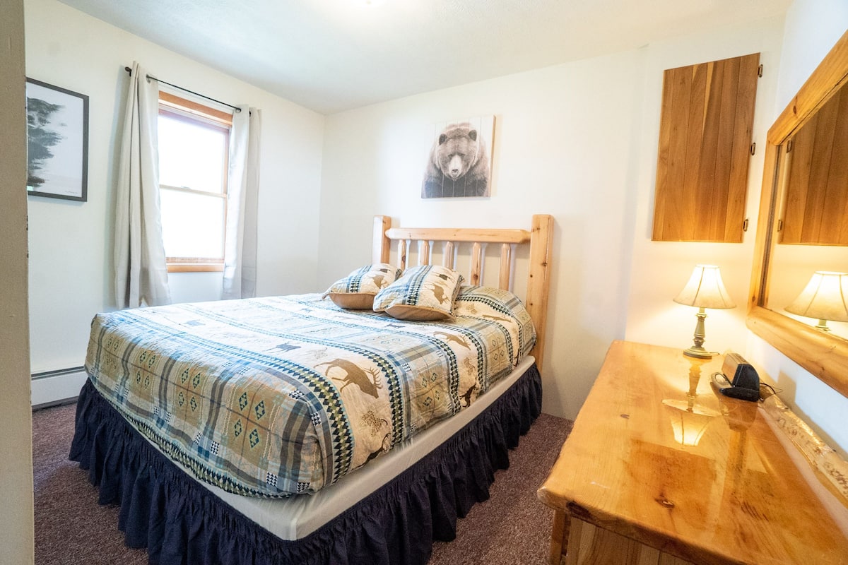 A bedroom with a bed, a wooden nightstand, a lamp, a window with curtains, and a bear-themed wall art.