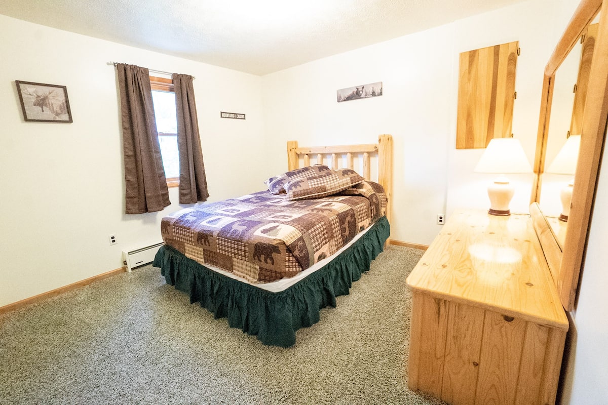 A bedroom with a wooden headboard, a bed with a patterned bedspread, a bedside table with a lamp, and a mirror.